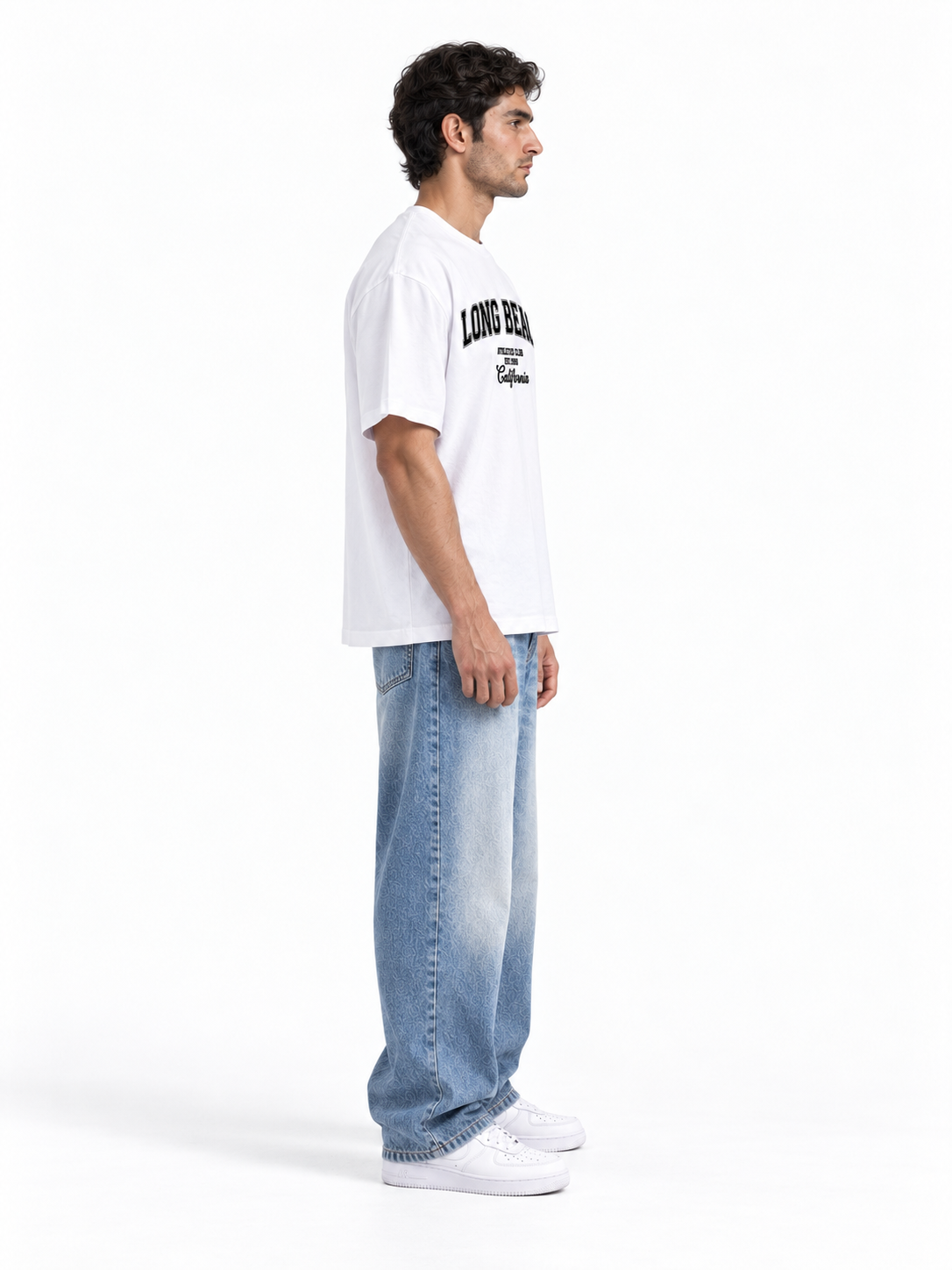 A man in profile wearing a white crewneck t-shirt with black collegiate-style text on the chest, paired with light-wash blue jeans and white sneakers, standing against a plain white studio background.