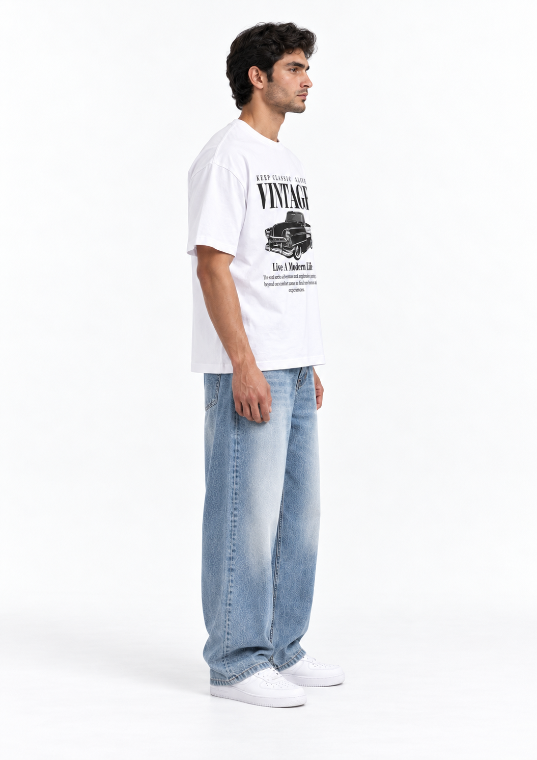 A man wearing a white vintage graphic t-shirt, light wash wide-leg jeans, and white sneakers, shown in profile against a plain white studio background.