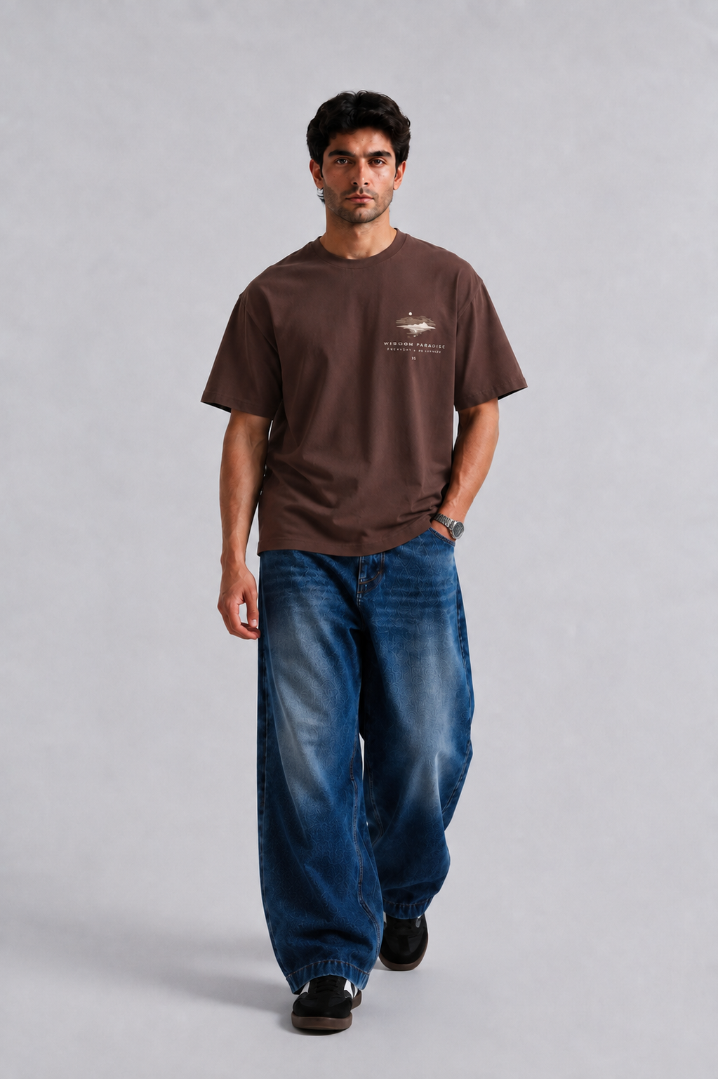 A man wearing a brown short-sleeve t-shirt with a small graphic on the left chest and loose-fit blue jeans, walking against a light gray studio background.