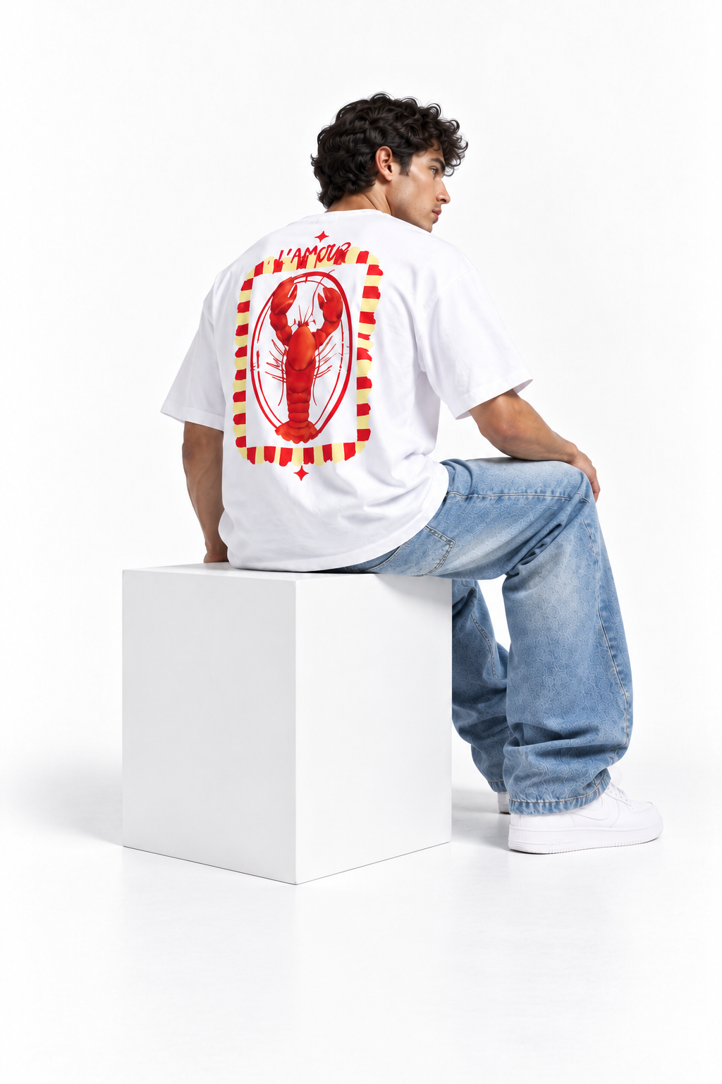 A man wearing a white t-shirt with a large red lobster graphic on the back, paired with light blue jeans and white sneakers, sitting on a white block against a white studio background.