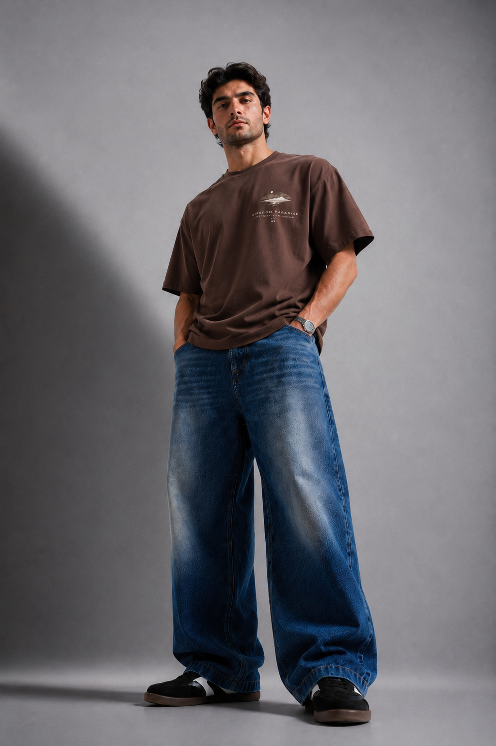 A man modeling a brown short-sleeve t-shirt with a small graphic on the left chest, paired with wide-leg blue jeans and black sneakers, standing against a gray studio background.