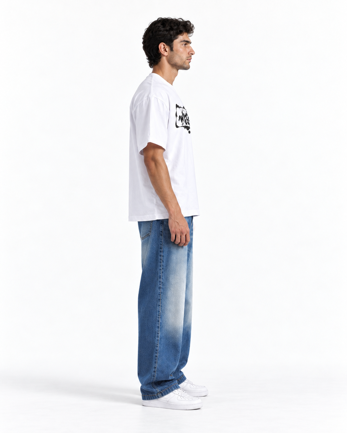 A man shown in profile wearing a white short-sleeve T-shirt with a black graphic print on the chest, paired with wide-leg faded blue jeans and white sneakers, against a plain white studio background.