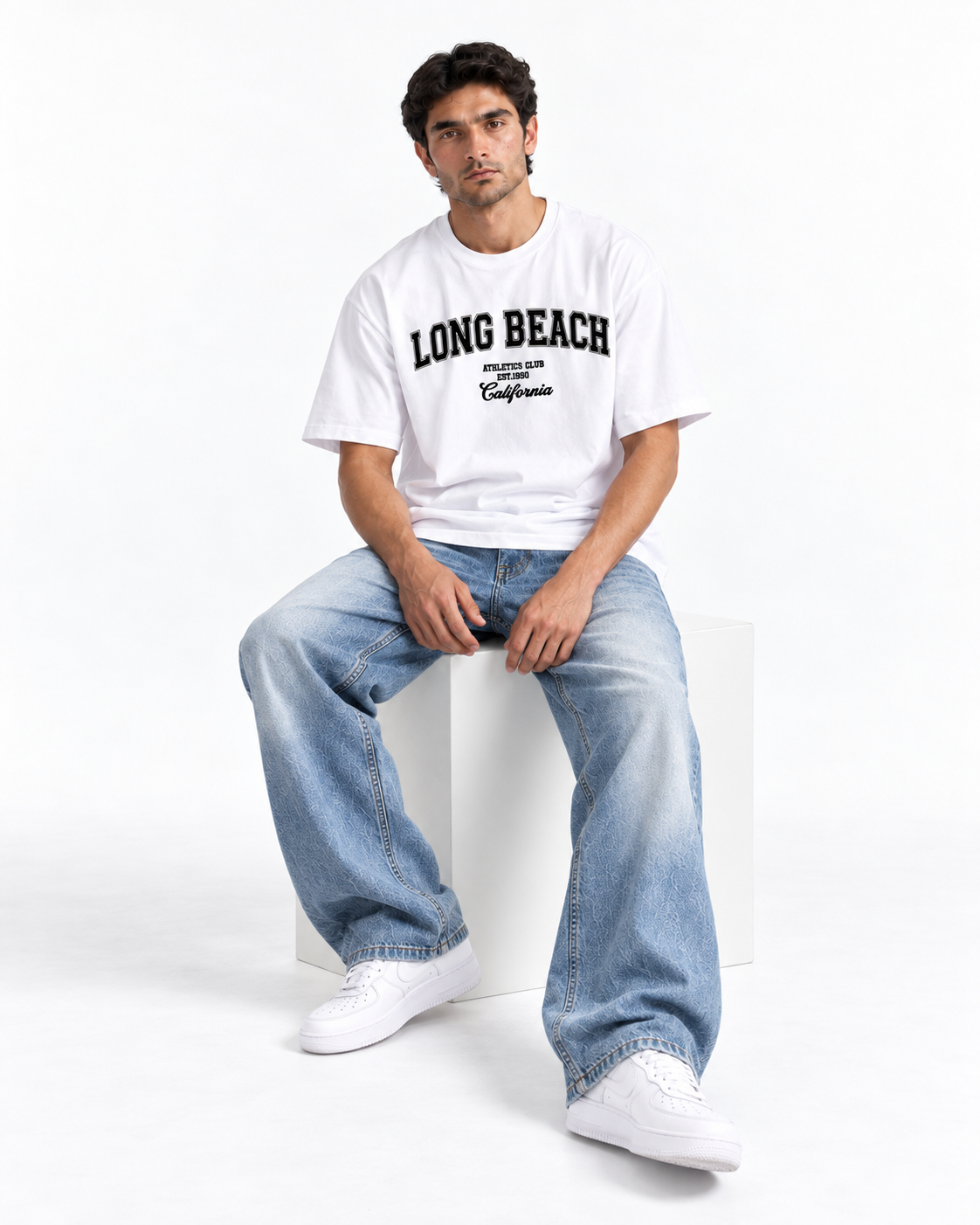 A man wearing a white crewneck t-shirt with 'LONG BEACH' printed in black on the chest, paired with light-wash blue jeans and white sneakers, sitting on a white block against a white studio background.