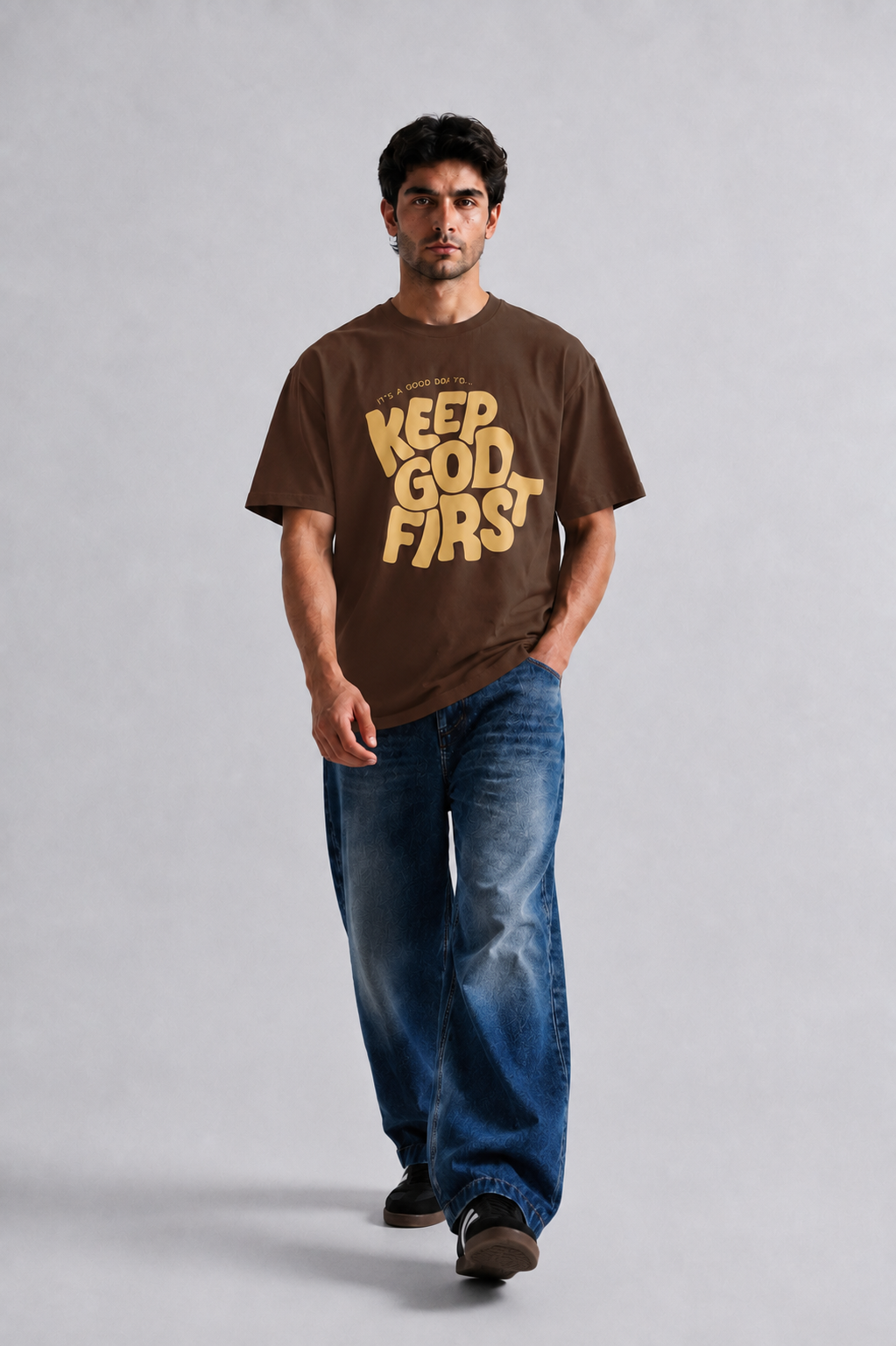 A man wearing a brown graphic t-shirt with 'KEEP GOD FIRST' printed in yellow, paired with blue jeans, walking against a light gray studio background.