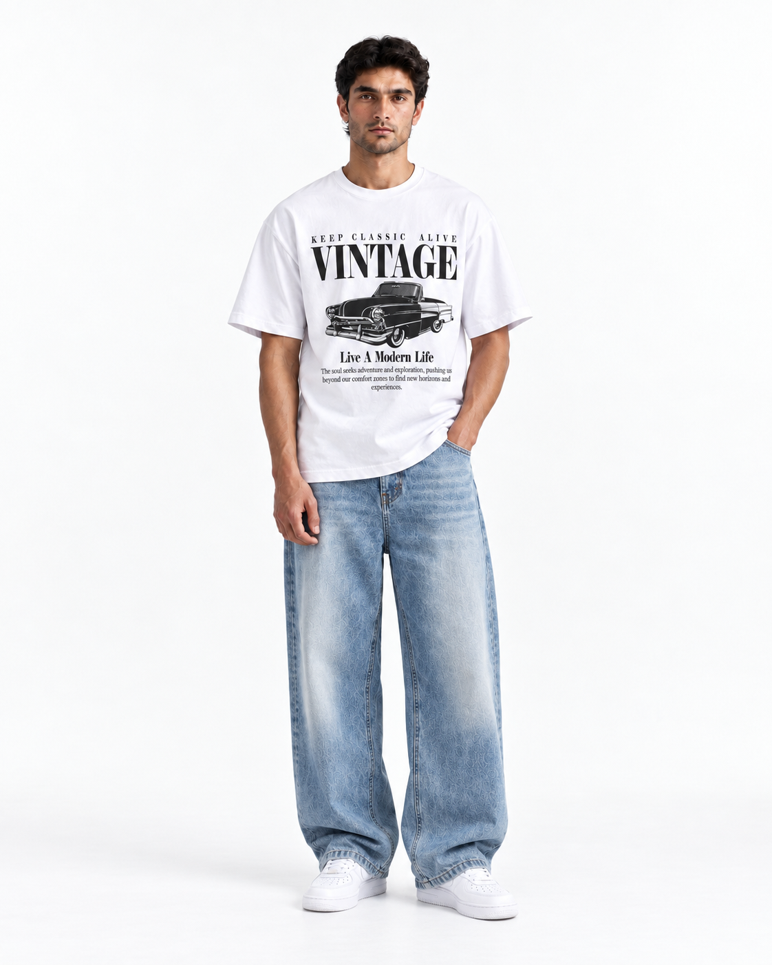 A man modeling a white vintage graphic t-shirt with a black car print and text, paired with wide-leg light blue jeans and white sneakers, against a plain white studio background.