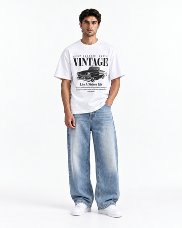 A man modeling a white vintage graphic t-shirt with a black car print and text, paired with wide-leg light blue jeans and white sneakers, against a plain white studio background.