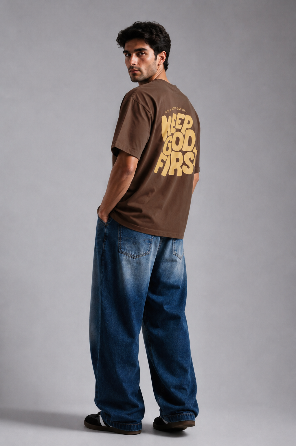A man wearing a brown t-shirt with a yellow graphic on the back and loose blue jeans, standing against a gray studio background.