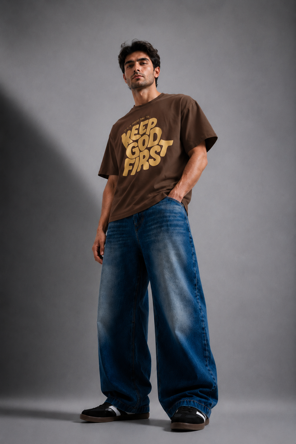A man modeling a brown oversized graphic t-shirt with 'KEEP GOD FIRST' printed in gold, paired with wide-leg blue jeans and black sneakers, against a dark gray studio background.
