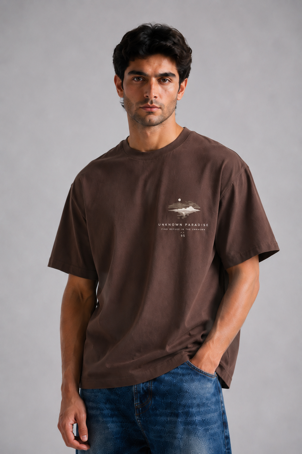 A man wearing a brown short-sleeve t-shirt with a small graphic on the left chest and blue jeans, standing against a neutral gray studio background.