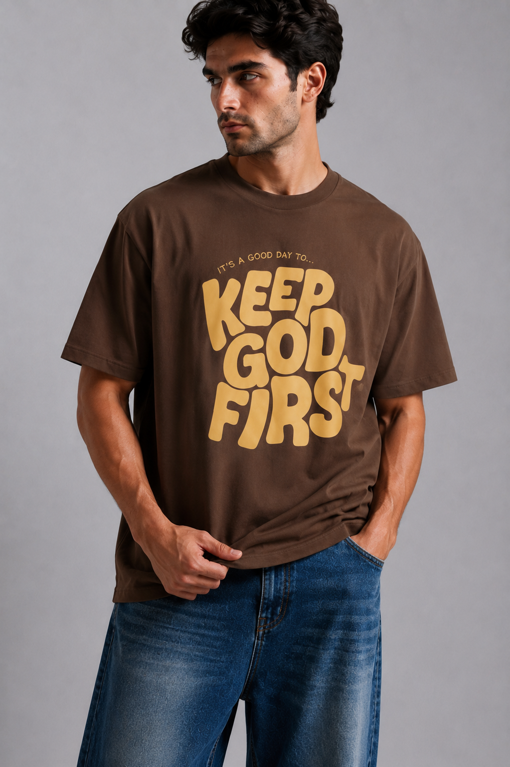 A man wearing a brown graphic t-shirt with the text 'KEEP GOD FIRST' in yellow, paired with blue jeans, against a solid gray studio background.