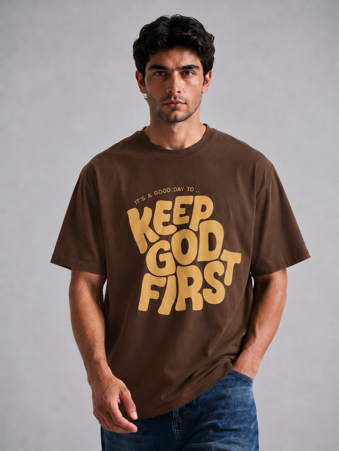 A man wearing a brown short-sleeve t-shirt with a large yellow graphic that reads 'KEEP GOD FIRST' and 'IT'S A GOOD DAY TO', paired with blue jeans, against a neutral gray background.