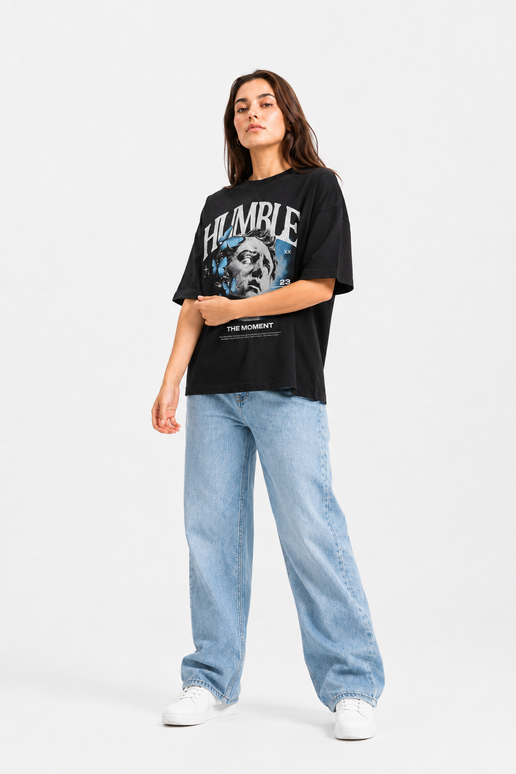 A woman modeling a black graphic t-shirt with a blue and white 'HUMBLE' design, paired with light-wash wide-leg jeans and white sneakers, against a plain white studio background.
