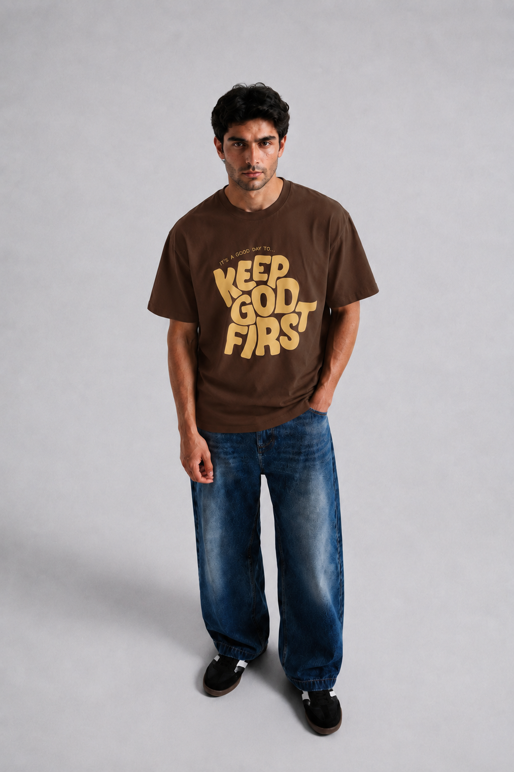 A man wearing a brown graphic t-shirt with 'KEEP GOD FIRST' printed in yellow on the front, paired with blue jeans and black shoes, standing against a light gray studio background.