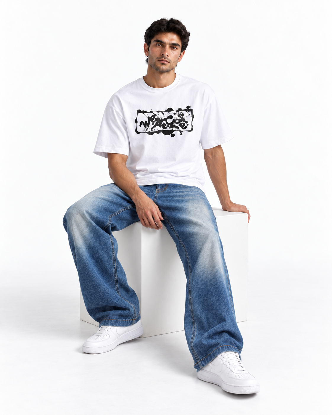 A man wearing a white crewneck t-shirt with a black graphic print, paired with wide-leg faded blue jeans and white sneakers, sitting on a white block against a white studio background.